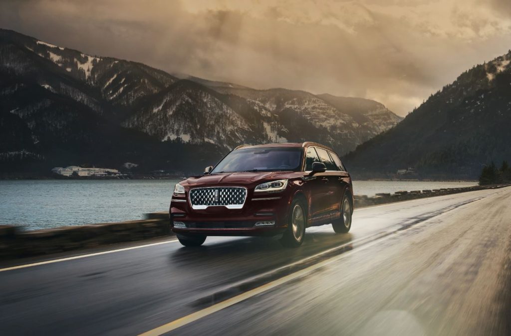 A red 2023 Lincoln Aviator driving down a mountain road. The sky is foggy and the sun is shining behind the clouds, the roads are wet.