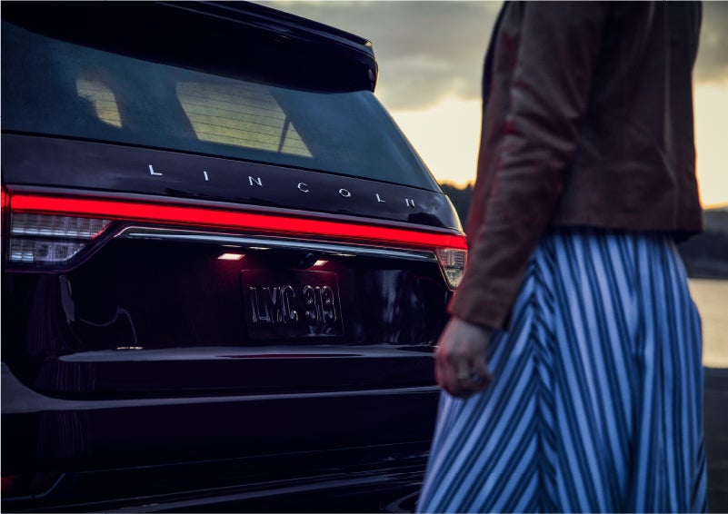 A person is shown near the rear of a 2023 Lincoln Aviator® SUV as the Lincoln Embrace illuminates the rear lights | Star Lincoln in Southfield MI