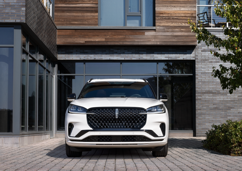 A white 2025 Lincoln Aviator® SUV is shown parked outside a modern building with gray brick and wood accents. | Star Lincoln in Southfield MI